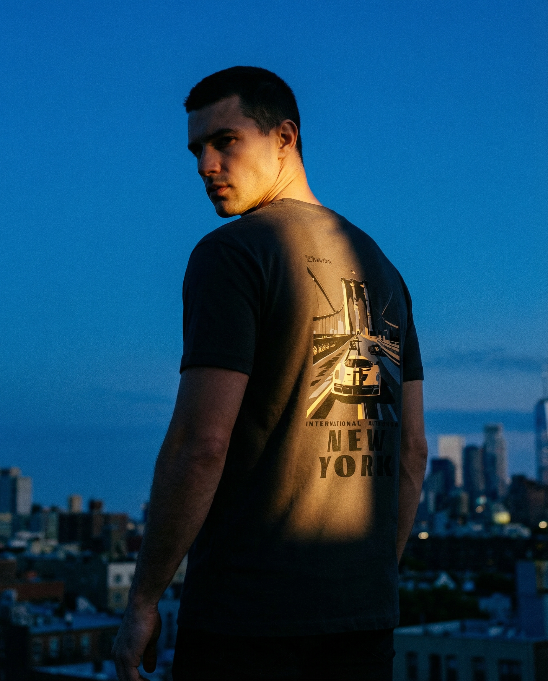 Man wearing a t-shirt with a New York design, standing against a city skyline at dusk.